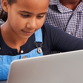 A child in a blue overall focuses on a laptop, seated with an adult in a checkered shirt, possibly assisting, in an indoor setting.