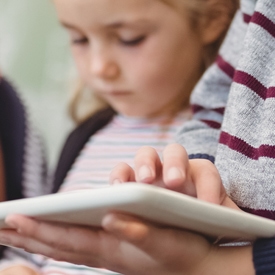 A child holds a white tablet, using it with one hand, while another young child in the background observes intently. The setting appears casual and indoors.