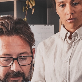 A man with glasses reads a document while a woman in a white shirt observes, in an office setting with shelves, a calendar, and a plant in the background.