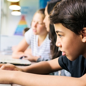 A young person focuses intently on a computer screen, seated in a classroom setting with other students working on computers nearby. Bright lighting and educational posters enhance the learning atmosphere.