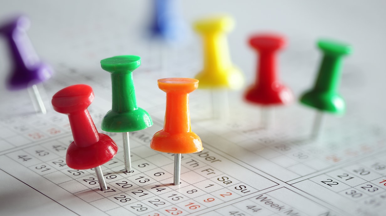 Colorful push pins are standing upright on a calendar, with dates clearly visible. The pins vary in color, including red, green, orange, yellow, and blue, indicating different events or reminders.