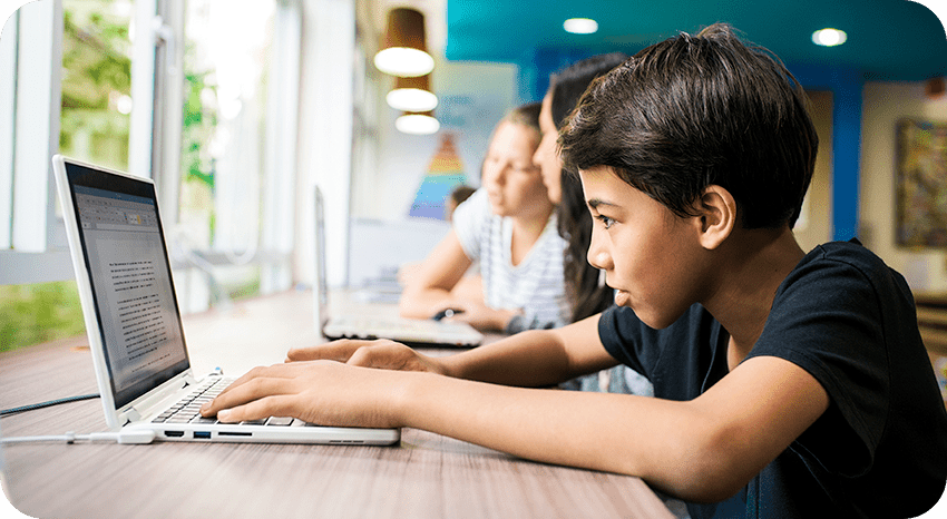 A focused child types on a laptop at a long table, with two other children working on laptops in the background, inside a bright room with large windows and blue walls.