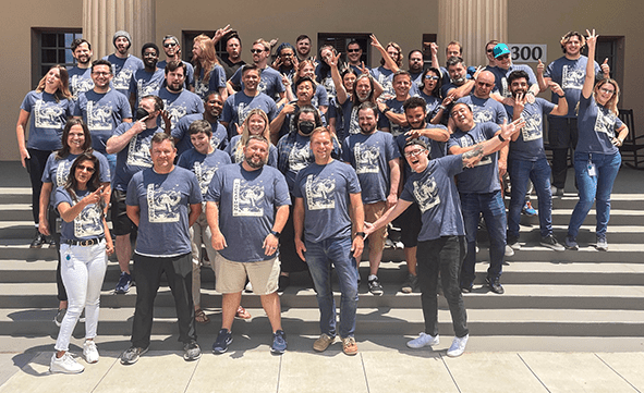 A large group of people wearing matching blue t-shirts pose energetically on steps outside a building. The building has columns and the number "300" is visible on a sign.
