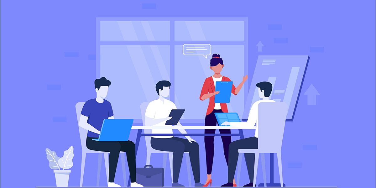 Four people are seated at a table with laptops and tablets, while one person stands presenting with a paper. A potted plant is nearby, and a window and screen are in the background.