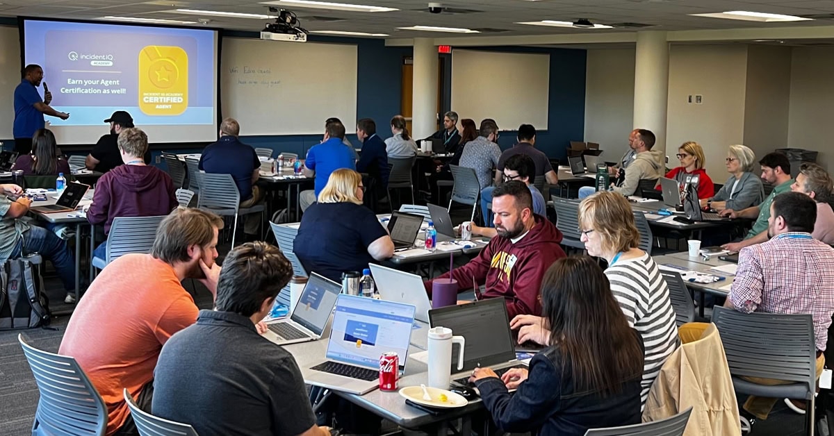Participants sit in a classroom, using laptops while a presenter points at a screen displaying, “Earn your Agent Certification as well! INCIDENT IQ ACADEMY. INCIDENT IQ CERTIFIED AGENT.”