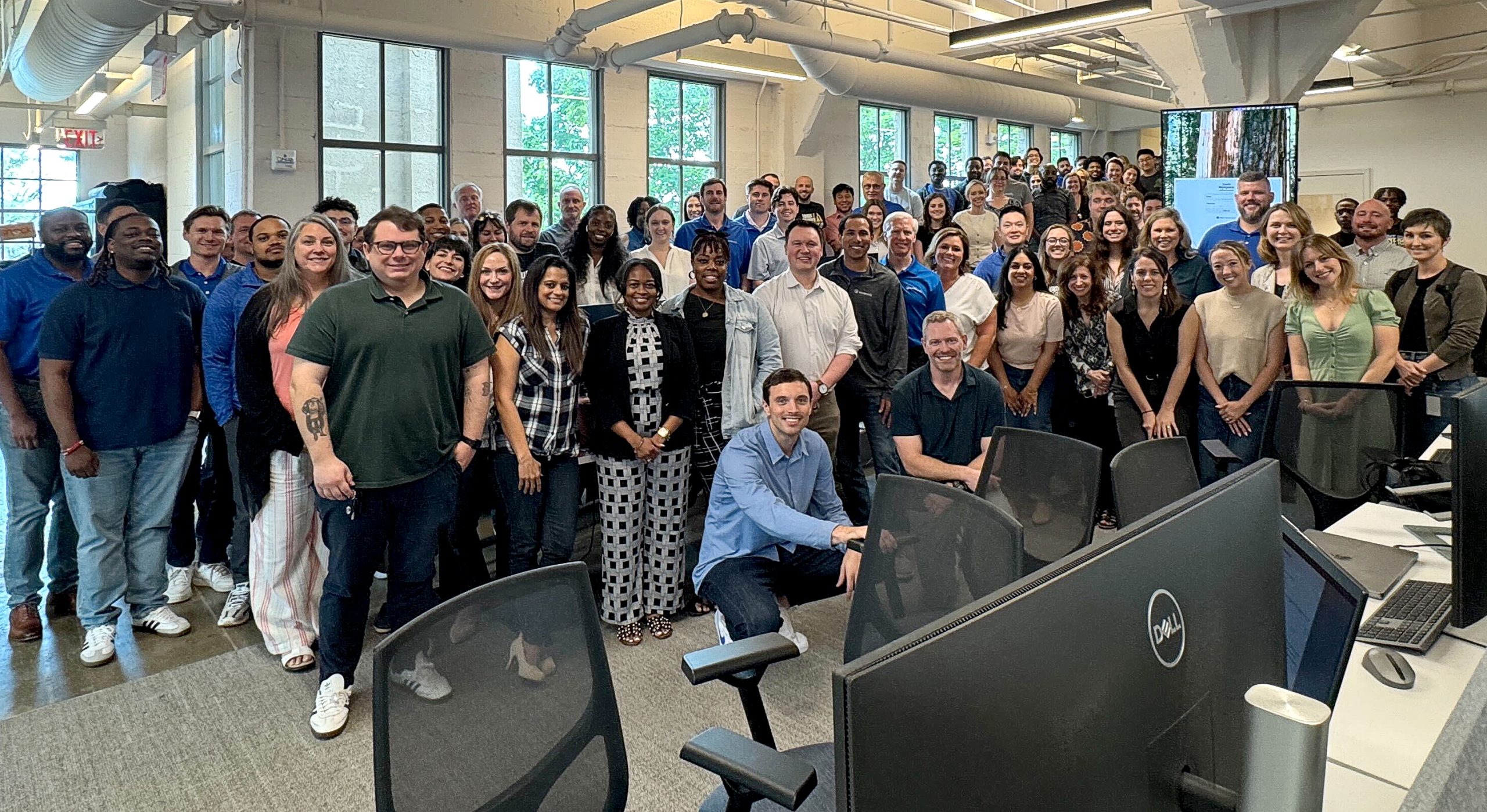 A large group of people gathers and smiles in an office setting with windows and computers, suggesting a workplace event or team meeting.