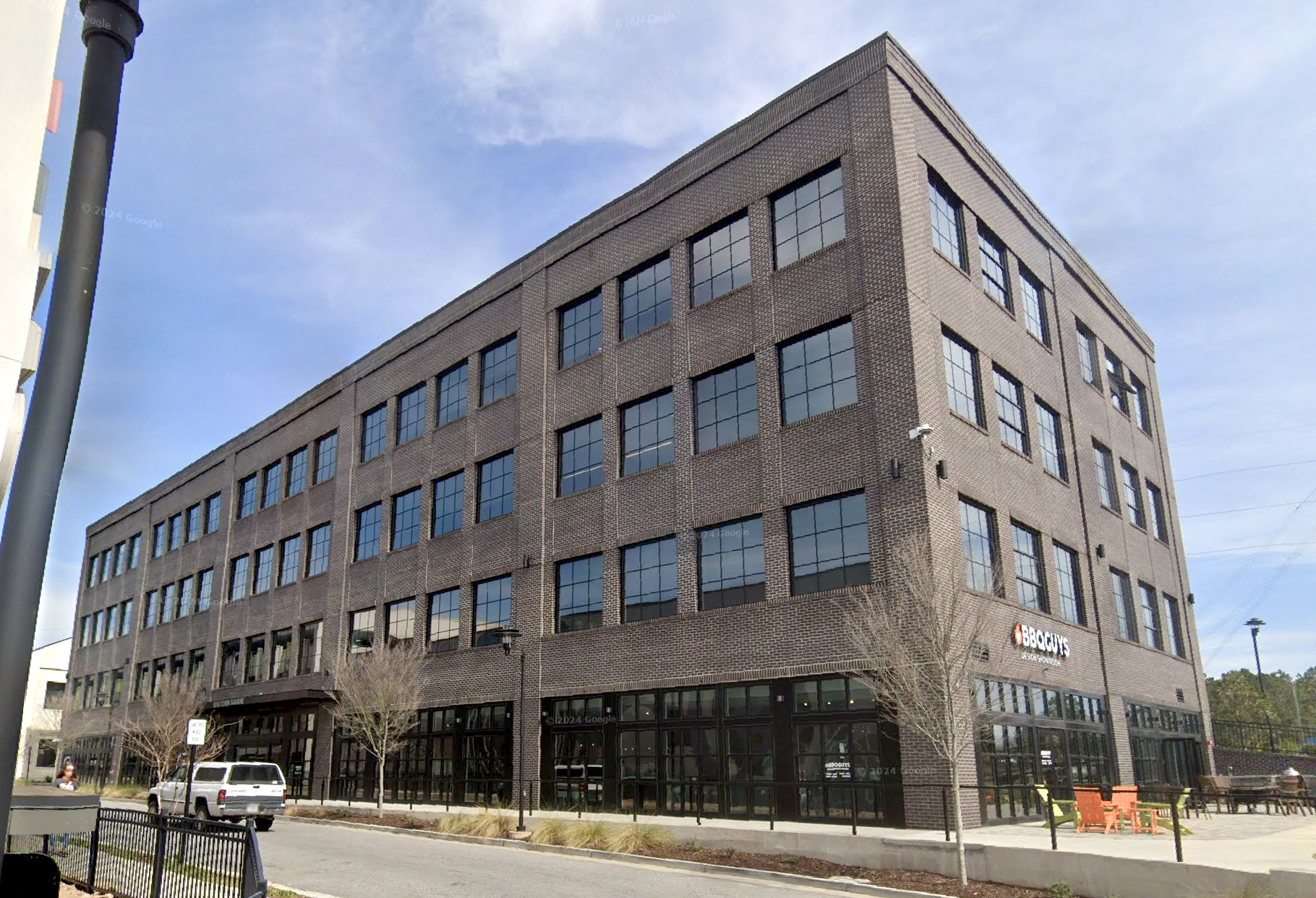A four-story brick building stands on a street corner with large framed windows. A sign reads “BBQGuys” above ground-level windows. Leafless trees and outdoor seating are visible nearby.